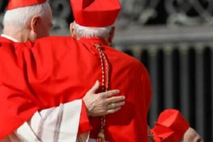 Dos cardenales vestidos de rojo, de pelo blanco, se abrazan en un consistorio