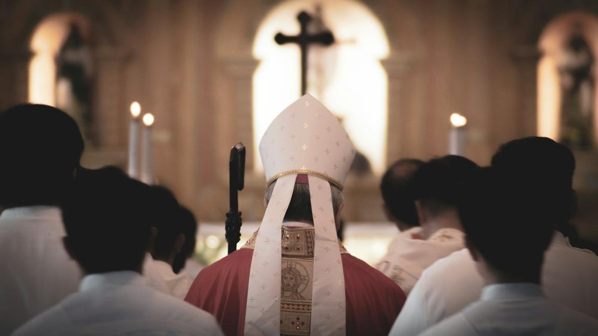 Un obispo rodeado de sacerdotes, vistos de espaldas en una iglesia.